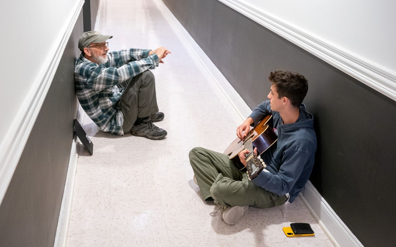 Older man sits and talks with younger boy in a hallway with a guitar