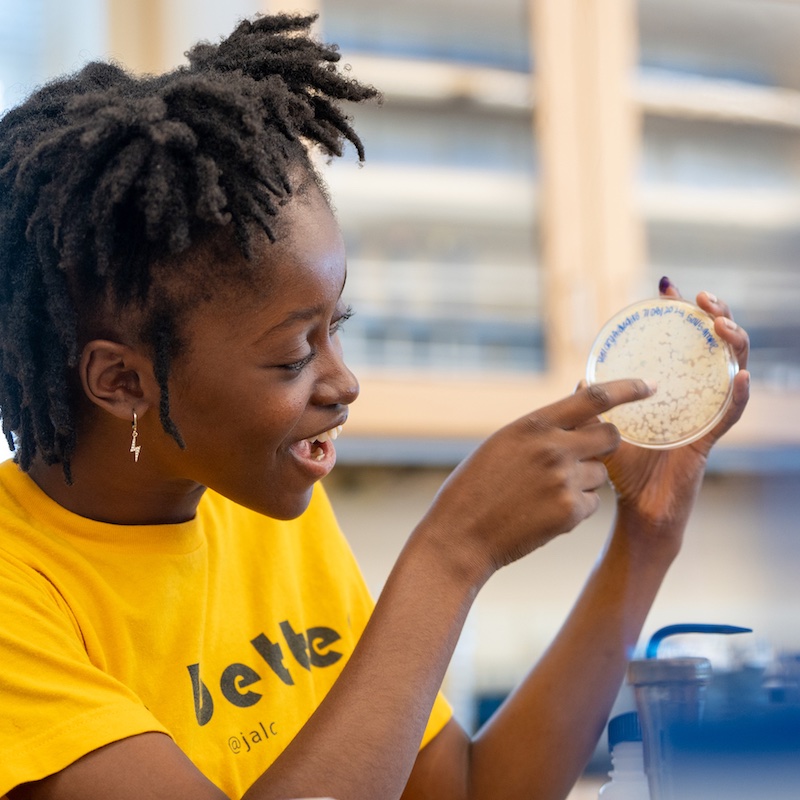 A high school student in a yellow shirt examines a petri dish.