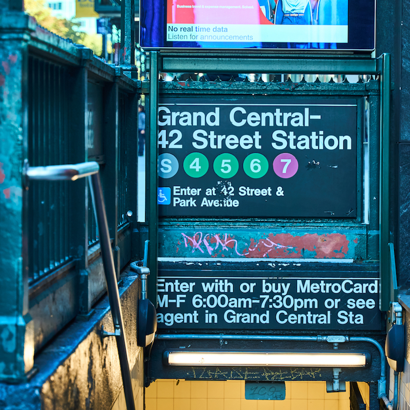 A New York City subway entrance sign for Grand Central-42 Street Station, featuring colored circles for lines S, 4, 5, 6, and 7. The sign includes accessibility information and operating hours (M-F 6:00am-7:30pm). The sign appears weathered with some graffiti, and has a blue-teal tint from the surrounding lighting. Above the entrance sign is a digital display showing "No real time data Listen for announcements"