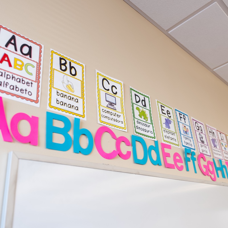 Classroom wall with colorful alphabet display. Large foam letters A-Z below bilingual English/Spanish alphabet flashcards showing letters and corresponding words with images.