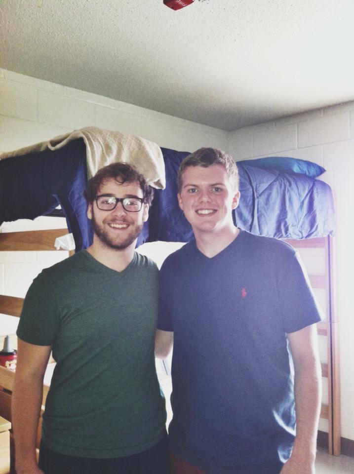 Two college students pose in a dorm room with bunk beds visible in the background. One wears glasses and a green t-shirt, the other wears a navy polo shirt.