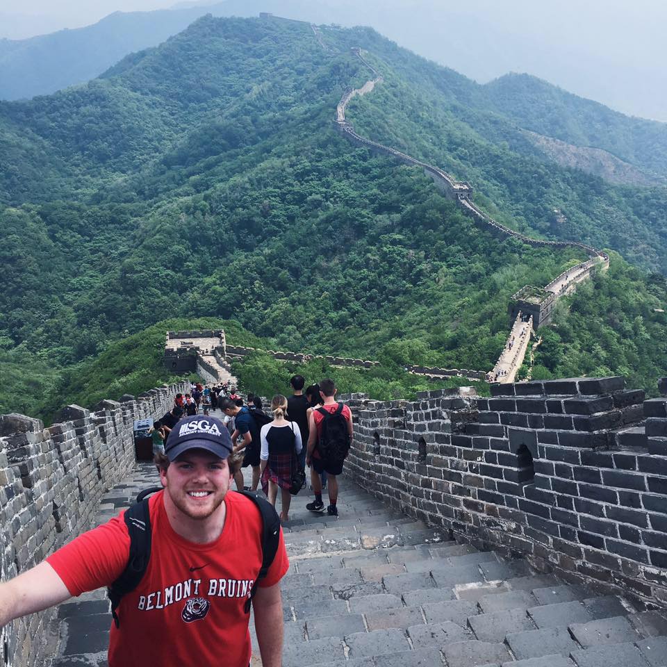 A person in a red Belmont Bruins t-shirt and baseball cap smiles at the camera while standing on the Great Wall of China. The wall stretches into the distance across green mountains.