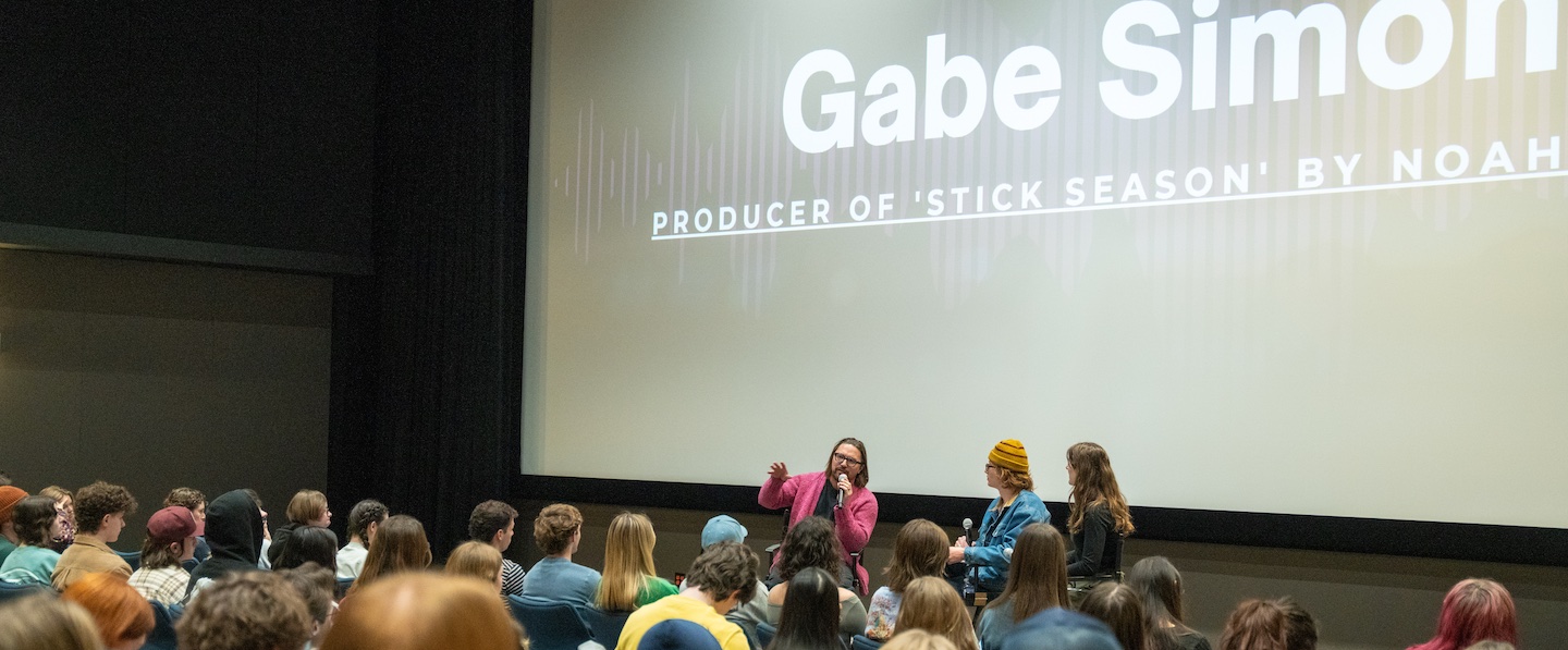 A presentation screen showing "Gabe Simon" with text "PRODUCER OF 'STICK SEASON' BY NOAH" visible below. In the foreground, a person with shoulder-length hair and wearing a pink cardigan (Gabe Simon) is speaking to an audience of students while holding a microphone. Two other people are standing nearby, including someone wearing a yellow beanie.