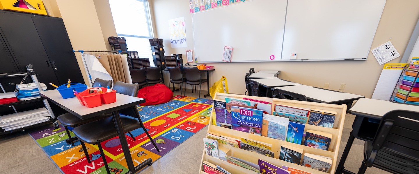 A classroom setting with a colorful alphabet learning station on the wall. There's a reading carpet with letters and numbers, student desks with red and blue supply bins, and a book display shelf containing children's literature. The room includes black chairs, white desks, and standard classroom storage cabinets.