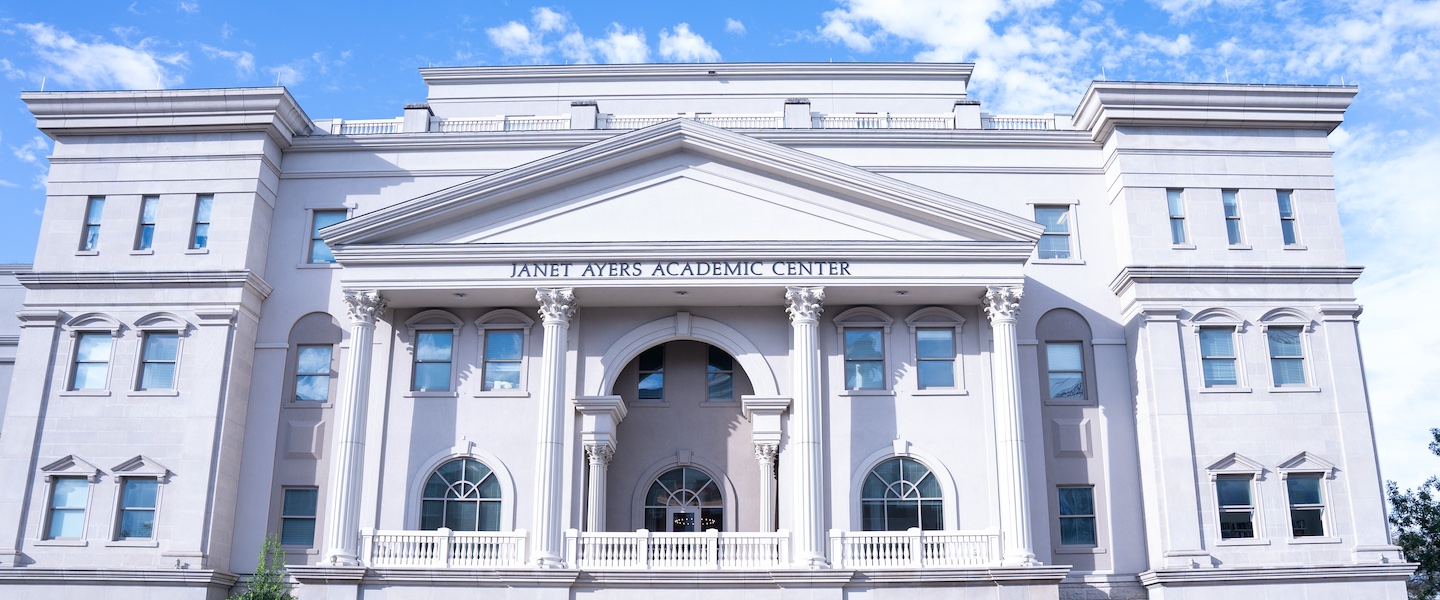 The Janet Ayers Academic Center at Belmont University features a grand neoclassical architectural design with a white facade, prominent Corinthian columns, and symmetrical wings. The three-story building showcases arched windows, decorative balustrades, and a triangular pediment above the main entrance where the center's name is displayed. The structure is photographed against a bright blue sky with white clouds.