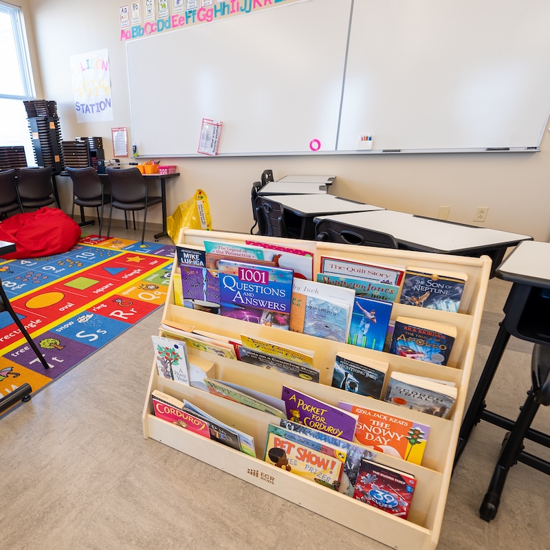 A classroom setting with a colorful alphabet learning station on the wall. There's a reading carpet with letters and numbers, student desks with red and blue supply bins, and a book display shelf containing children's literature. The room includes black chairs, white desks, and standard classroom storage cabinets.