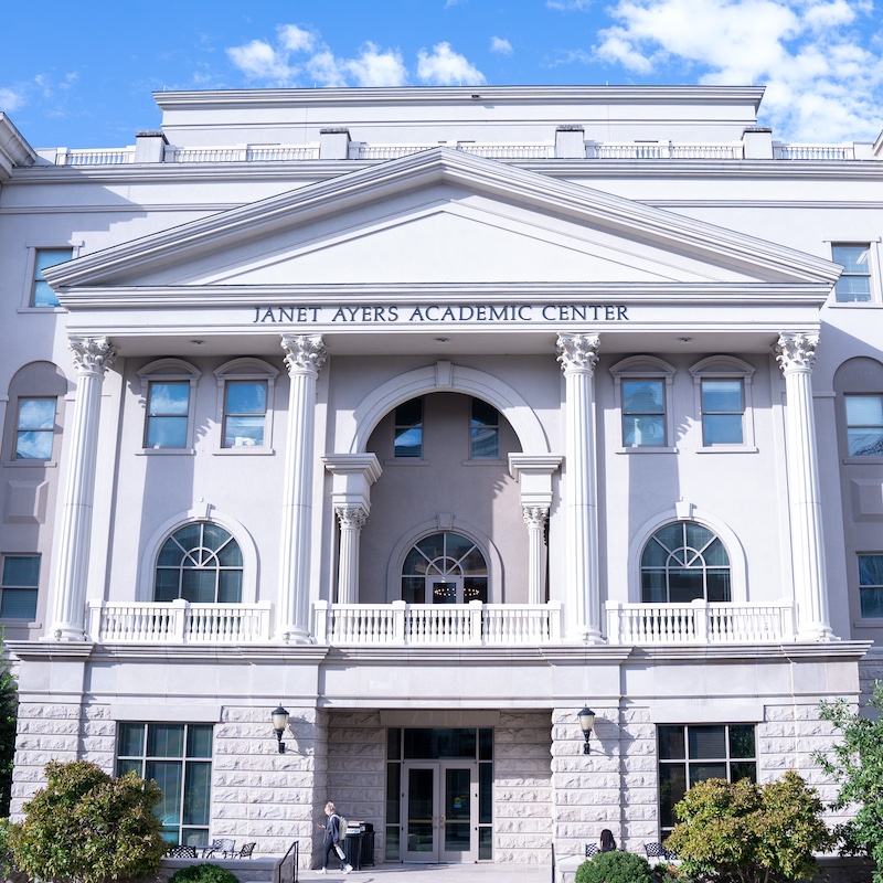 The Janet Ayers Academic Center at Belmont University features a grand neoclassical architectural design with a white facade, prominent Corinthian columns, and symmetrical wings. The three-story building showcases arched windows, decorative balustrades, and a triangular pediment above the main entrance where the center's name is displayed. The structure is photographed against a bright blue sky with white clouds.