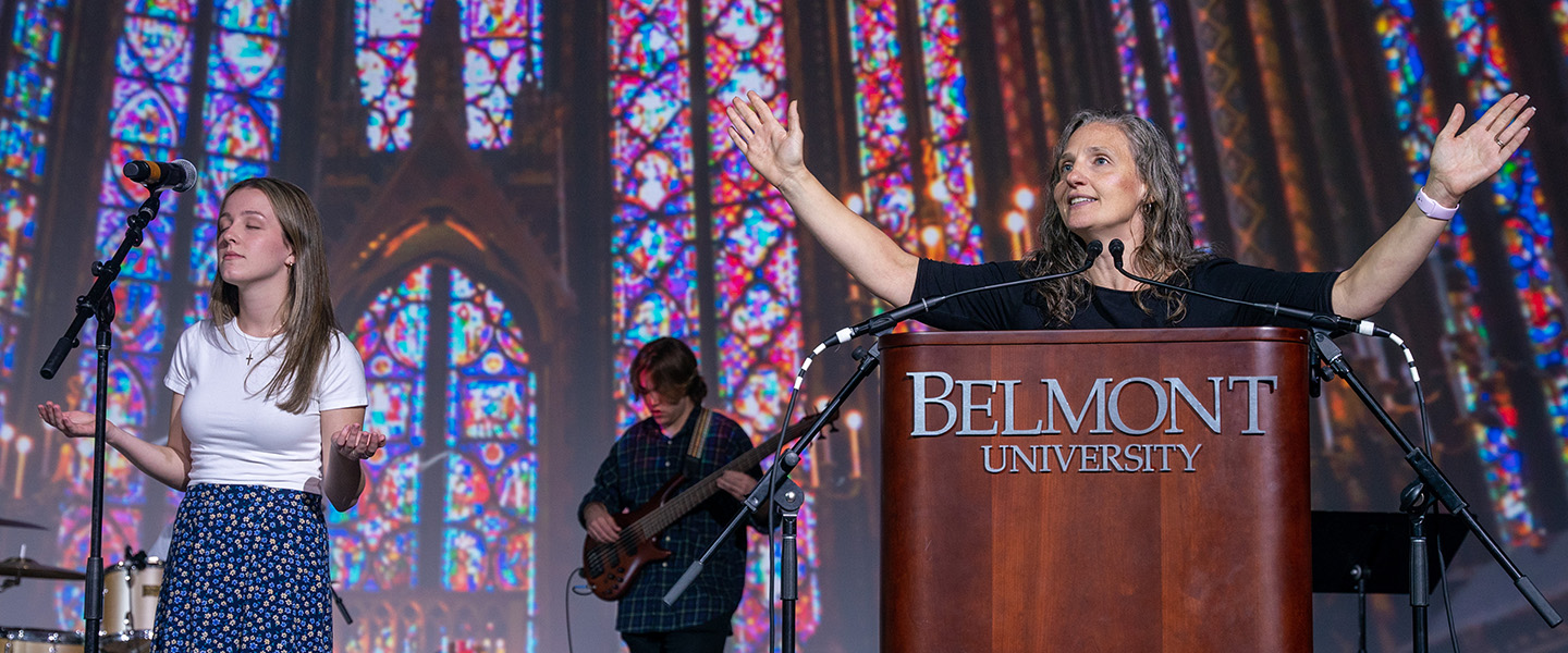 A woman stands behind a podium and addresses the chapel, raising her arms.