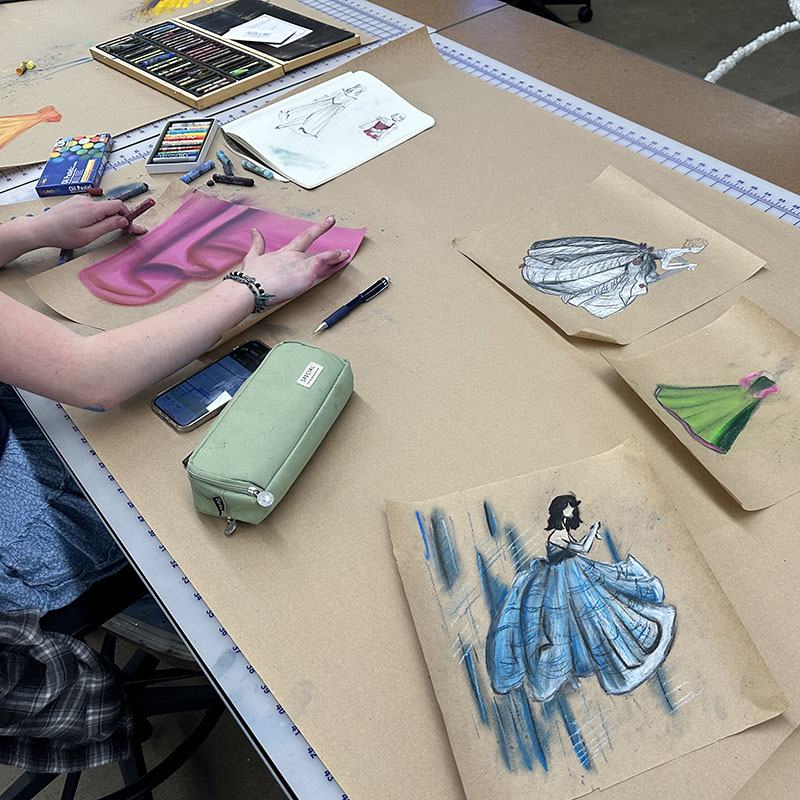 Student working with a pink pastel to create a design with other designs on paper surrounding her 