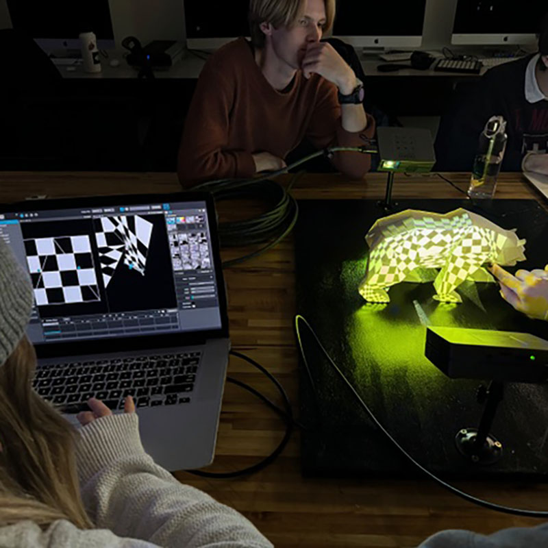 Two students work on an immersive project, one is on the computer and the other is working with a projection mapped animal bear model.