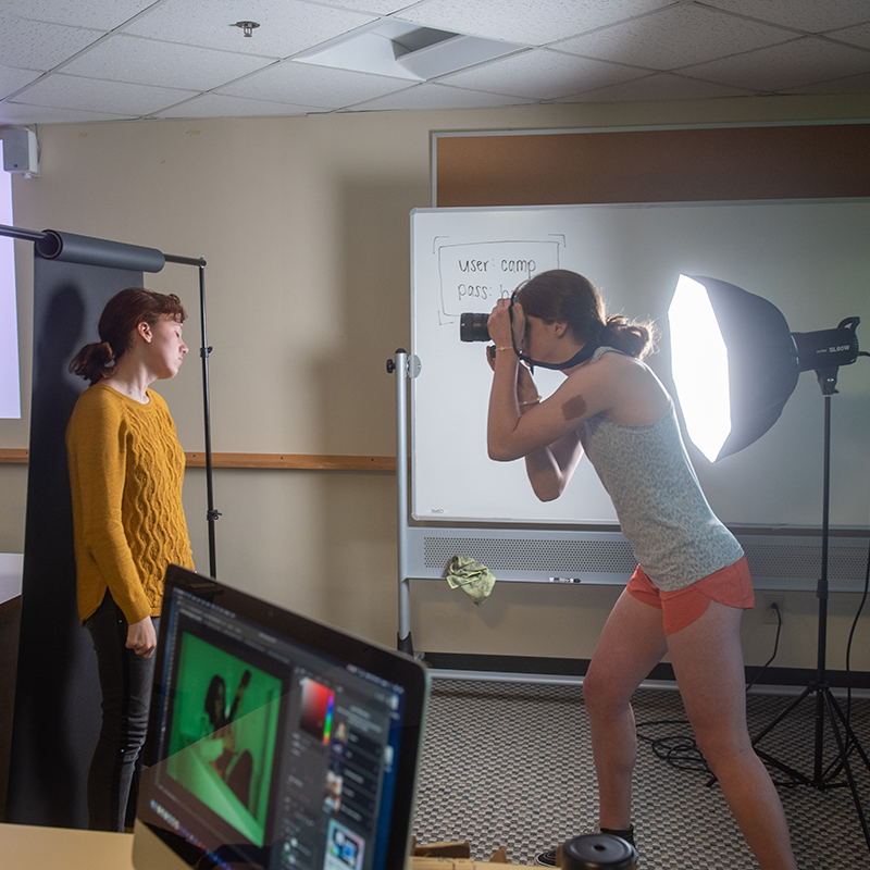 One student takes the photo of another student in front of the lighting equipment.